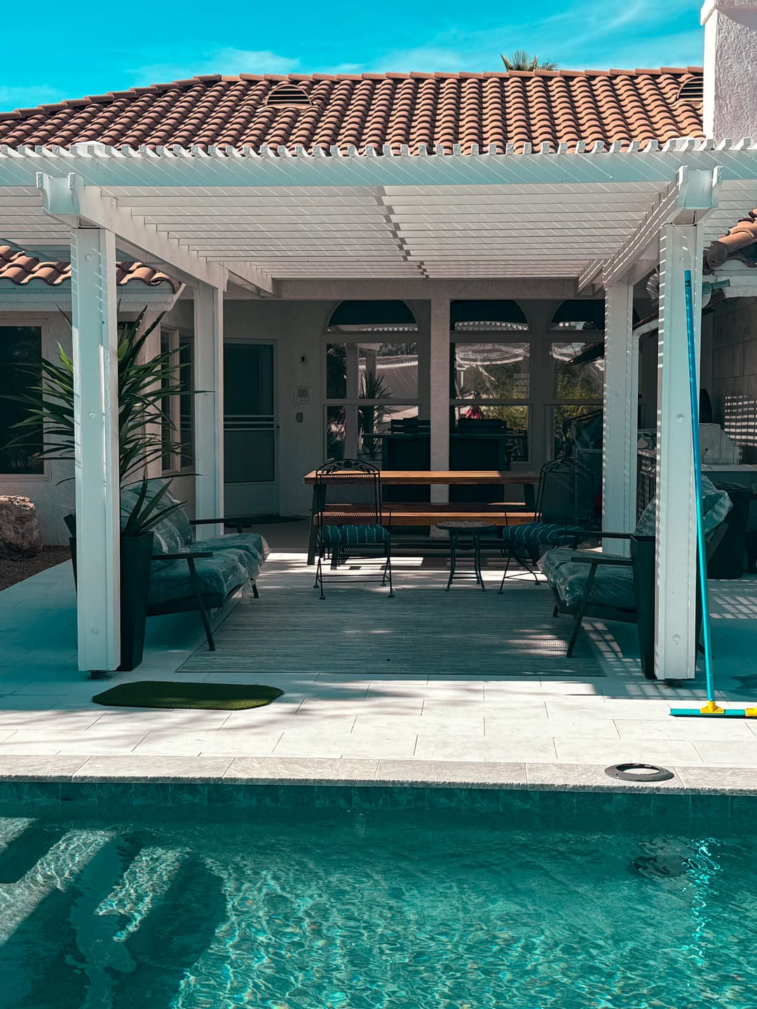 Sunny patio with pergola, table, chairs, and pool in a residential backyard setting.
