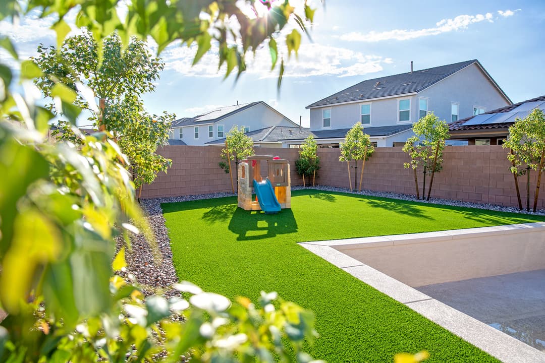 Bright backyard with artificial grass, playground slide, and trees under a clear blue sky.